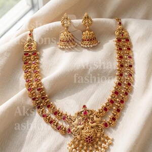 May include: A gold-toned necklace and earring set with red gemstone accents and small pearl drops. The necklace features a detailed pendant with a bird design. The earrings are bell-shaped with pearl accents. The jewelry is displayed on a white cloth.