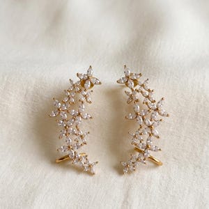 May include: A pair of gold-toned ear climbers featuring a cluster of marquise-cut clear cubic zirconia stones arranged in a floral-inspired pattern. Each earring has a curved metal wire frame designed to follow the contour of the earlobe.