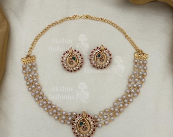May include: Gold-toned necklace and earring set. The necklace features three strands of faux pearls and a central pendant with red and green accents. Matching earrings complete the set, showcasing a peacock design. The brand name "Akshar fashions" is visible.
