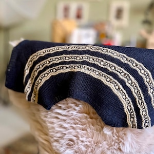 Op de afbeelding: Een marineblauwe gebreide sjaal met een decoratief, herhalend patroon in een lichtbeige kleur. De sjaal is gedrapeerd over een zacht, lichtbruin oppervlak, wat een gezellige en gestructureerde uitstraling creëert. Het ontwerp heeft meerdere rijen van het beige patroon.