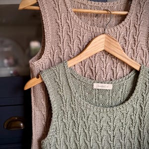 Op de afbeelding: Twee mouwloze tops met kabelbreisel op houten kleerhangers. Eén is stoffig roze van kleur en de andere is een gedempt groen. De tops hebben een textuur, geweven ontwerp en een ronde halslijn. Het label op de groene top luidt "loveknit.nl".