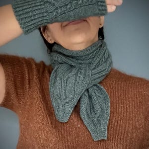 Op de afbeelding: Een gebreide teal sjaal en bijpassende vingerloze handschoenen. De sjaal is om de nek geknoopt en de handschoenen bedekken de ogen. Het breiwerk heeft een kabelpatroon. De persoon draagt een bruine trui.
