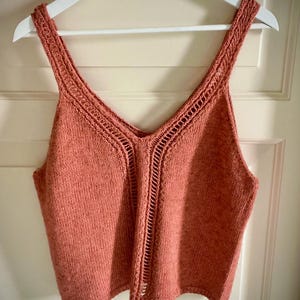 Puede incluir: Un top de punto de color coral con cuello en V y delicados detalles calados. La parte superior se muestra en una percha blanca, mostrando su tejido de punto texturizado y el diseño de las correas. El color de la prenda es un tono cálido y apagado.