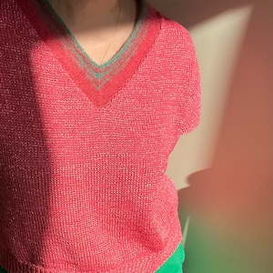 Pode incluir: Um suéter de malha rosa com uma borda verde e branca ao redor do decote. O suéter é feito de um material macio e leve e tem um caimento relaxado.