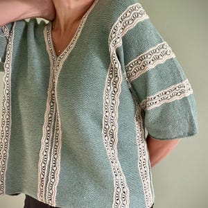 Op de afbeelding: Een zeegroene gebreide top met een V-hals en korte mouwen. De top heeft crèmekleurige kanten randen langs de randen en mouwen. De gebreide stof heeft een getextureerd uiterlijk.