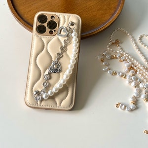 May include: A beige quilted phone case with a pearl chain and a silver charm with a heart design. The chain is attached to the case with a metal ring.