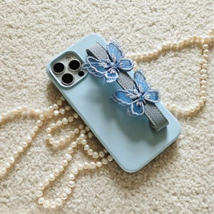 May include: A light blue phone case with a gray strap and two blue butterfly embellishments. The phone case is lying on a white fuzzy surface with a pearl necklace.