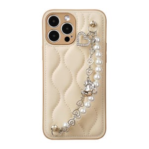 May include: Cream-colored phone case with a quilted pattern and gold-colored trim. The case features a decorative charm with a heart-shaped design and a pearl beaded strap. The phone case is designed to protect the phone.