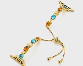 May include: Gold-toned Apple Watch band with a decorative design. The band features a series of oval-shaped, colorful gemstones in shades of blue and orange. The band has a chain-style adjustable closure.