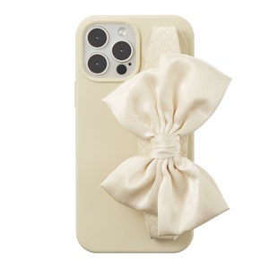 May include: A cream-colored phone case with a large cream-colored satin bow attached to the back. The case has a textured design and a camera bump with three lenses.