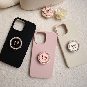 May include: Three phone cases with different colored backgrounds. The cases are black, pink, and beige. Each case has a pearl and gold bow ring holder.