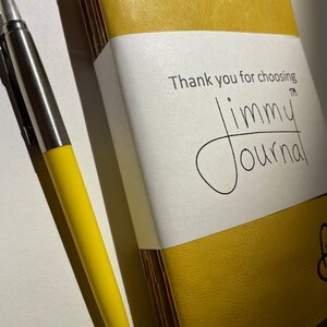 Pode incluir: Um diário encadernado em couro amarelo com o texto "Thank you for choosing jimmy journal™" na capa. Uma caneta amarela está ao lado do diário.
