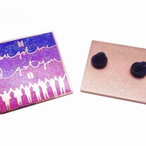 Könnte beinhalten: Eine quadratische Emaille-Anstecknadel mit einem glitzernden lila und rosa Farbverlauf als Hintergrund. Die Anstecknadel zeigt eine weiße Silhouette von sieben Personen, die sich an den Händen halten, mit dem Text "If you got me I got you" in weißer Kursivschrift.