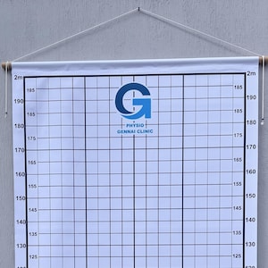 May include: A white wall-mounted height chart banner featuring a black grid and numerical markings from 65 to 200 cm. The top center displays a blue logo with the text G PHYSIO GENNAI CLINIC. The banner is suspended by a wooden rod and white cord.