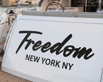 May include: The stern of a white yacht featuring a teak wood deck and a black script logo that reads Freedom with the text NEW YORK NY printed in a sans-serif font below it. The vessel includes a metal railing and a steering wheel visible above.