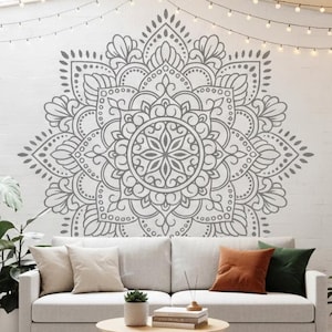 May include: A bright living room with a light grey sofa, cushions, and a round wooden coffee table. Potted plants are placed around the room. A large grey mandala design is on the white wall, and fairy lights hang from the ceiling.