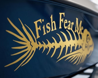 May include: A dark blue boat hull featuring a gold-colored decal of a fish skeleton graphic. The text "Fish Fear Me" is printed in a gold-colored serif font above the skeletal fish design, which spans horizontally across the curved surface.