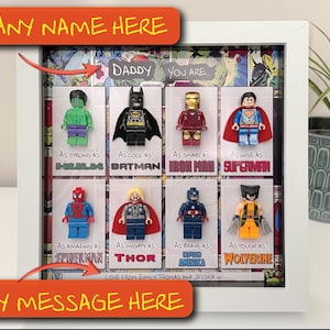 Może przedstawiać: Oprawione dzieło sztuki z figurkami superbohaterów Lego, w tym Hulka, Batmana, Iron Mana i Supermana. Grafika zawiera tekst "Daddy You Are" oraz obszary tekstowe do personalizacji imienia i wiadomości. Rama jest biała.