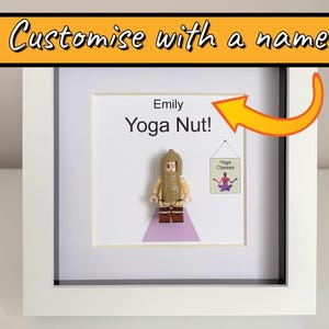 Op de afbeelding: Een ingelijst kunstwerk met een Lego-figuur in een yogahouding, met de tekst "Emily Yoga Nut!" en een klein bordje met "Yoga Classes". De lijst is wit en het kunstwerk staat tegen een witte achtergrond. De bovenkant van de lijst heeft de tekst "Customise with a name".
