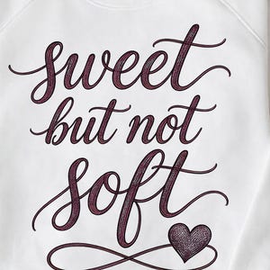May include: White sweatshirt with the phrase "Sweet but not Soft" in a dark purple script. A heart graphic is below the text. The sweatshirt is folded on a woven surface.