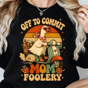 May include: A black crewneck sweatshirt featuring a graphic of a white duck wearing heart-shaped sunglasses and a bandana, riding a teal and orange scooter. The text reads OFF TO COMMIT MOM FOOLERY, surrounded by retro sunset and flower motifs.