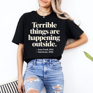 Könnte beinhalten: Schwarzes T-Shirt mit dem Zitat "Terrible things are happening outside." in fetter, cremefarbener Schrift. Das Zitat wird Anne Frank, 1944, und Amerikanern, 2026, zugeschrieben. Das Shirt wird mit einer blauen Jeans kombiniert.