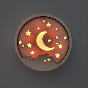 Puede incluir: Una luz de noche de madera con forma de luna creciente con estrellas y nubes. La luna está iluminada con una luz amarilla cálida.