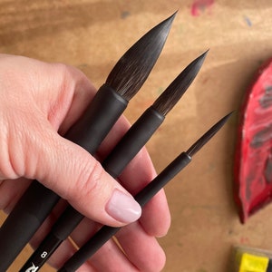 Könnte beinhalten: Drei schwarze Aquarellpinsel mit dunklen Borsten. Die Pinsel werden in einer Hand gehalten, wobei die Spitzen der Pinsel nach oben zeigen. Die Pinsel sind unterschiedlich groß, der größte Pinsel links und der kleinste rechts. Der Hintergrund ist eine hellbraune Oberfläche.