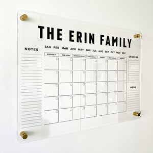 May include: A clear acrylic wall calendar featuring the text THE ERIN FAMILY at the top. It includes a monthly grid with days of the week, months of the year, a notes section, a grocery list, and a menu section, all secured by four gold-toned standoffs.
