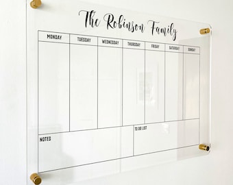 May include: A clear acrylic weekly wall planner featuring black printed text and grid lines. The header reads The Robinson Family, followed by columns for Monday through Sunday, and bottom sections labeled Notes and To Do List.