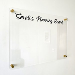 May include: A rectangular clear acrylic planning board featuring the black text Sarah's Planning Board printed on the surface. The board is mounted to the wall with four circular gold-toned metal standoffs located at each of the four corners.