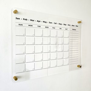 May include: A clear acrylic monthly calendar with black printed text and gold-colored mounting hardware. The calendar grid displays days of the week and months of the year, with a notes section on the right side. The calendar is mounted on a white wall.