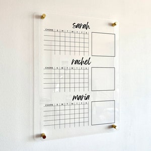 May include: A clear acrylic chore chart featuring three sections labeled with the names Sarah, Rachel, and Maria. Each section includes a grid for days of the week and a blank square, mounted to the wall with four gold-toned metal standoffs.