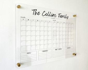 May include: A clear acrylic wall calendar featuring the text The Collins Family, two monthly grids, and sections labelled Notes, Reminders, What's Cooking, and Groceries. The board is mounted with four gold-toned circular hardware standoffs.