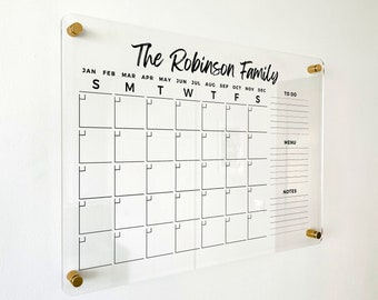 May include: Clear acrylic family calendar with gold-colored hardware. The calendar is labeled "The Robinson Family" and includes a monthly grid, a to-do list, a menu section, and a notes section. The calendar is mounted on a white wall.