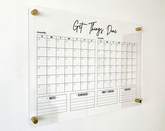 May include: A clear acrylic wall calendar featuring the text Get Things Done at the top. It includes two monthly grids with columns for days of the week, plus sections labelled Notes, Reminders, What's Cooking, and Groceries. Four gold-toned metal standoffs secure the board to the wall.