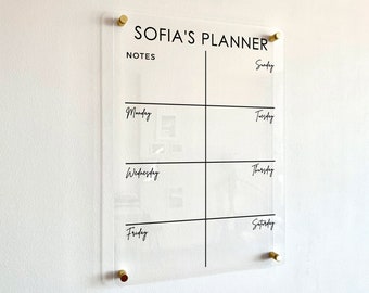 Może przedstawiać: Przezroczysty akrylowy planer ścienny ze złotymi elementami montażowymi. Tablica zawiera u góry napis SOFIA'S PLANNER, sekcję na notatki oraz tygodniowy harmonogram z dniami tygodnia nadrukowanymi czarną czcionką.