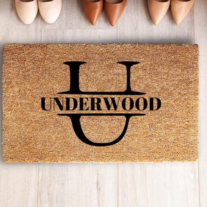 May include: A rectangular tan coir doormat featuring a large black letter U with the name UNDERWOOD printed in black capital letters across the center of the letter. The mat has a coarse, fibrous texture and a clean, simple design.