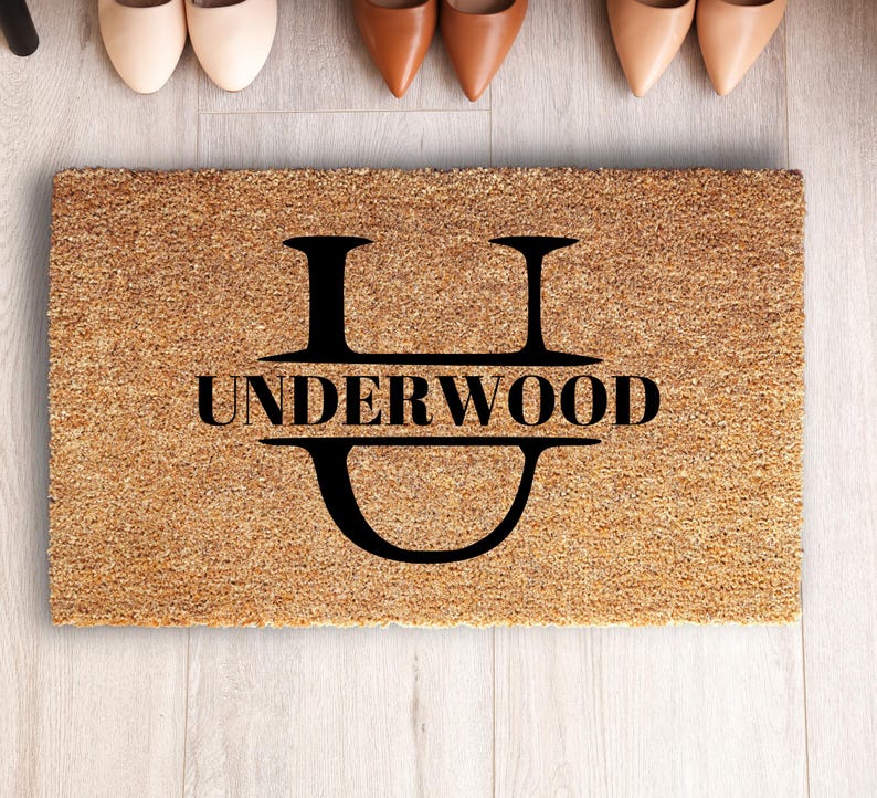 May include: A rectangular tan coir doormat featuring a large black letter U with the name UNDERWOOD printed in black capital letters across the center of the letter. The mat has a coarse, fibrous texture and a clean, simple design.