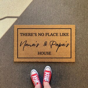 May include: A rectangular doormat with the text "THERE'S NO PLACE LIKE Nana's & Papa's HOUSE" in black lettering. The mat is brown and has a black border. Red sneakers with white laces are visible at the bottom.