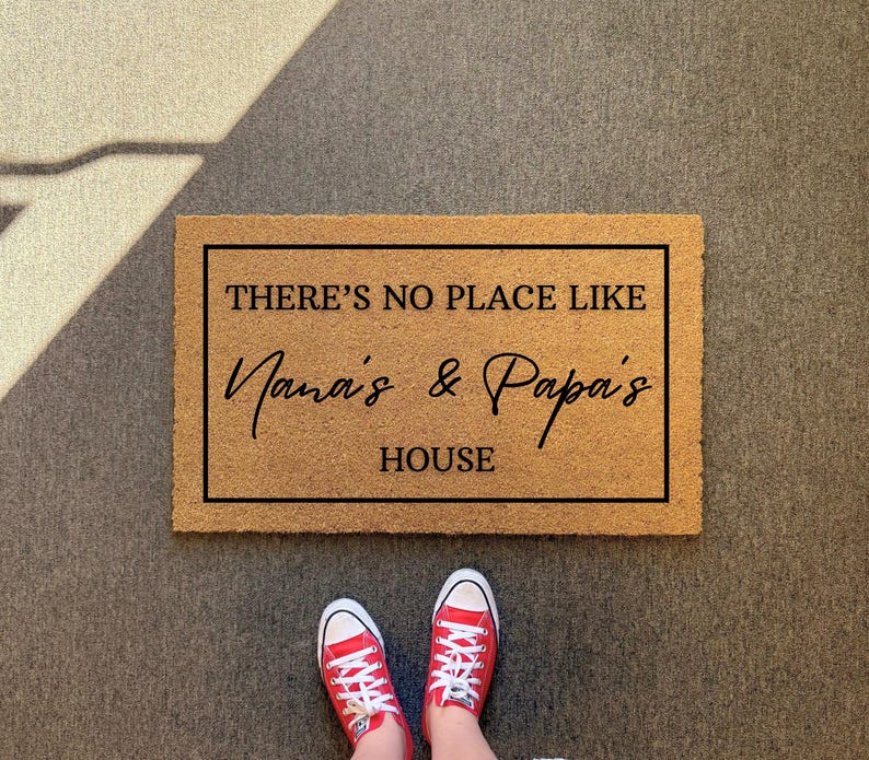 May include: A rectangular doormat with the text "THERE'S NO PLACE LIKE Nana's & Papa's HOUSE" in black lettering. The mat is brown and has a black border. Red sneakers with white laces are visible at the bottom.