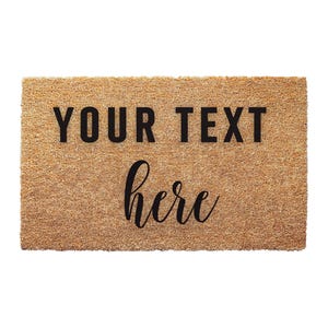 May include: A rectangular tan coir doormat with a coarse, fibrous texture. The mat features black printed text in two different fonts. The top line reads YOUR TEXT in a bold, sans-serif typeface, and the bottom line reads here in a script font.