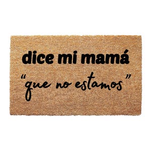 May include: Rectangular doormat made of natural coir fiber. The mat features the phrase "dice mi mamá 'que no estamos'" in black, bold lettering. The doormat is a light brown color.