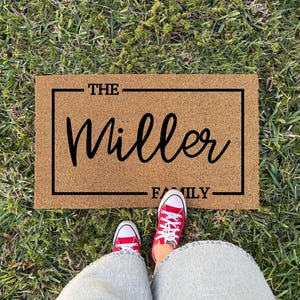 May include: A rectangular tan coir doormat featuring a black rectangular border. The text THE is printed in black above the word Miller in a large cursive script, with the word FAMILY printed in black below, all centered within the border.