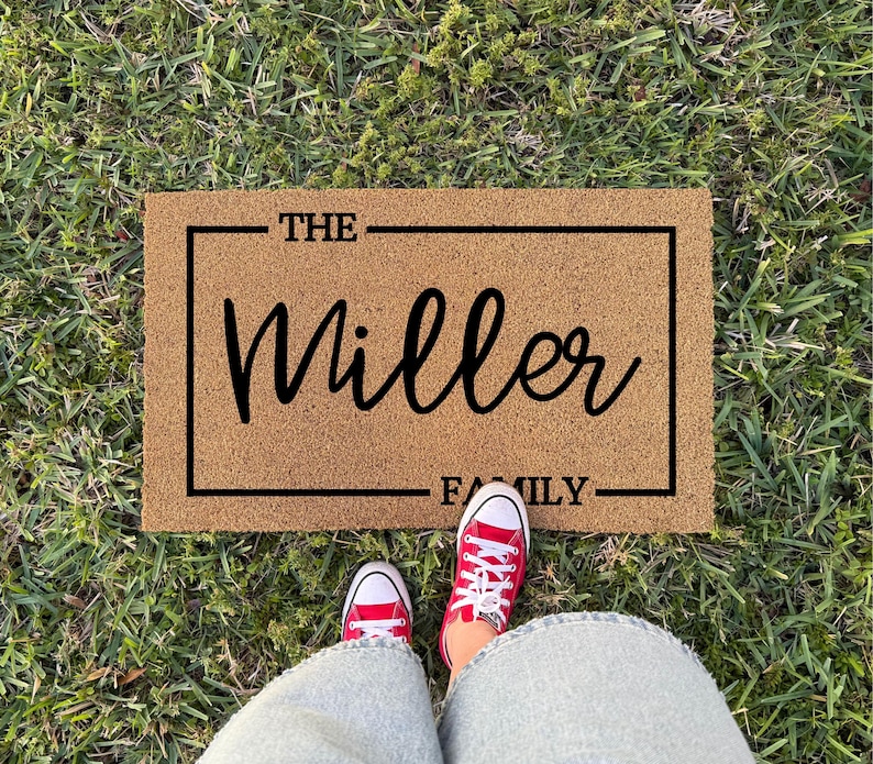 May include: A rectangular tan coir doormat featuring a black rectangular border. The text THE is printed in black above the word Miller in a large cursive script, with the word FAMILY printed in black below, all centered within the border.