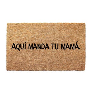 May include: A rectangular doormat made of natural coir fiber. The mat features the phrase "AQUÍ MANDA TU MAMÁ" in bold, black letters. The text is centered on the mat, which has a light brown color.