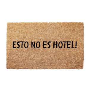 May include: Rectangular doormat made of natural coir fibers. The mat features the phrase "ESTO NO ES HOTEL!" in bold, black capital letters, set against a light brown background. The doormat is designed for outdoor use.