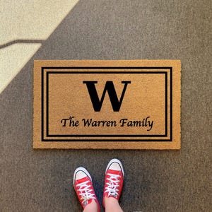 May include: A rectangular tan coir doormat featuring a black double-line border, a large black letter W in the center, and the text The Warren Family printed in black below the letter. The mat has a coarse, natural fiber texture throughout.