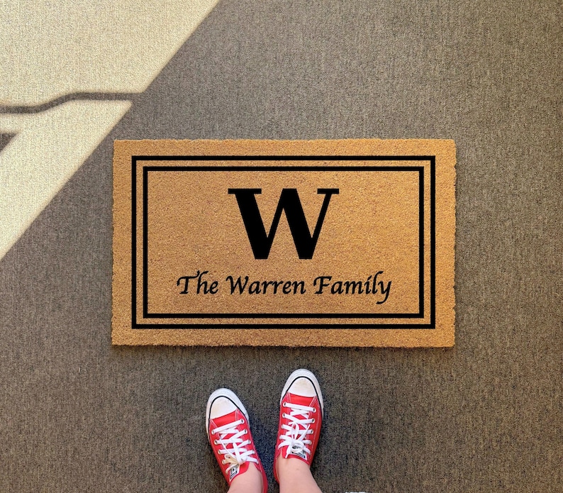 May include: A rectangular tan coir doormat featuring a black double-line border, a large black letter W in the center, and the text The Warren Family printed in black below the letter. The mat has a coarse, natural fiber texture throughout.