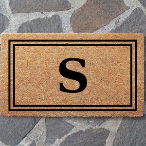 May include: A rectangular tan coir doormat featuring a bold black letter S centered within a double-lined black rectangular border. The natural fiber mat has a coarse, textured surface and a clean, geometric design printed in black ink.